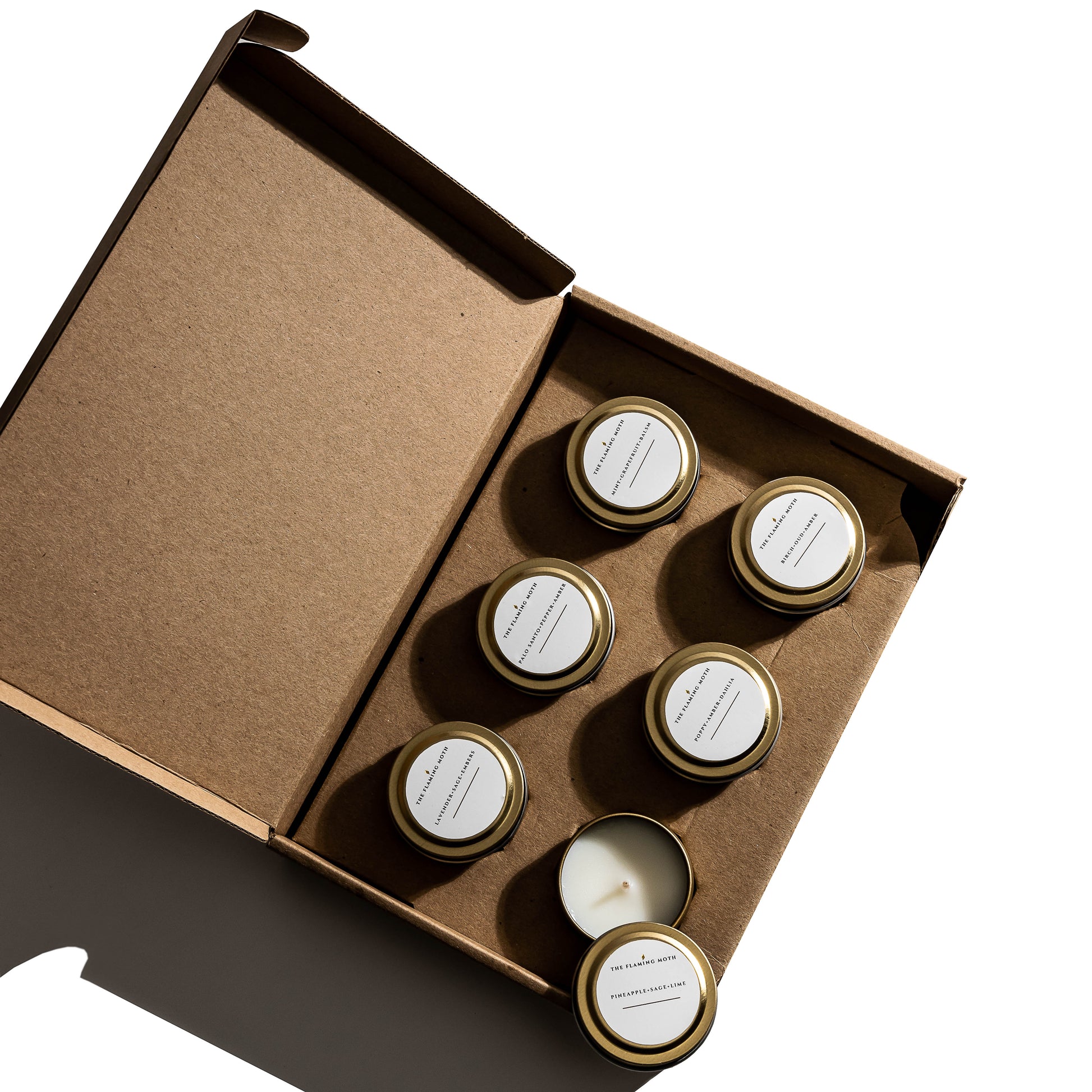 Set of six small candles in jars with gold lids in a cardboard box on a white background