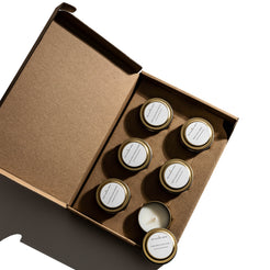 Set of six small candles in jars with gold lids in a cardboard box on a white background